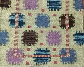 May include: A beige rug with a repeating pattern of squares and rectangles in shades of blue, brown, and purple. The rug has a fringed edge and a geometric design. The rug is made of woven material.