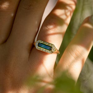 Könnte beinhalten: Ein goldener Ring mit einem rechteckigen, teal-farbenen Edelstein in der Mitte, umgeben von kleineren, klaren Steinen. Der Ring hat ein geometrisches Design, vor einem unscharfen Hintergrund.