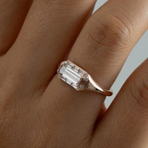 Könnte beinhalten: Ein roségoldener Ring mit einem rechteckigen, klaren Edelstein mit geometrischem Design. Der Ring hat ein poliertes Metallband, und der Edelstein ist in einer detaillierten, eckigen Lünette gefasst.