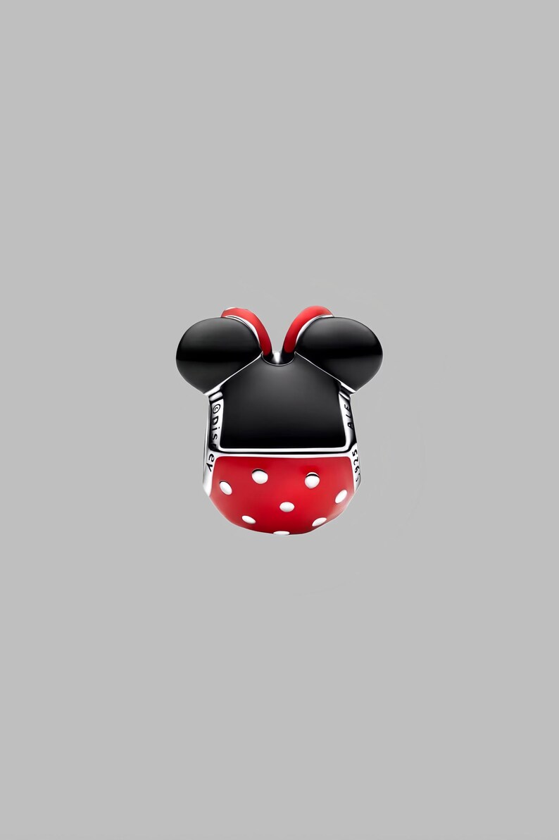 Puede incluir: Un charm con forma de cabeza de Minnie Mouse. El charm presenta orejas negras, una falda roja con lunares blancos y una parte superior negra. El charm es de metal y tiene la palabra "Disney" grabada en el lateral.