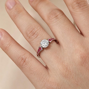 May include: A silver-toned ring featuring a large, round, clear center stone held in a six-prong setting. The band is accented with a row of small, round, vibrant red stones embedded along each side of the central setting, creating a contrast.