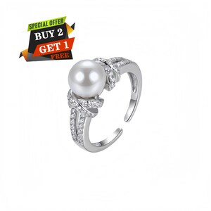 Puede incluir: Un anillo abierto de tono plateado con una perla blanca central y pequeñas piedras transparentes incrustadas a lo largo de la banda. Un gráfico en la esquina superior izquierda muestra el texto SPECIAL OFFER BUY 2 GET 1 FREE en blanco, rojo y negro.