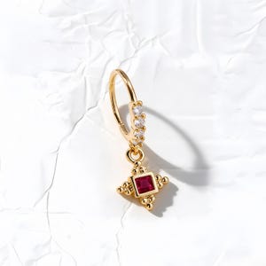 May include: A gold hoop earring featuring a row of small clear stones along the upper band and a dangling square charm set with a single deep red stone, accented by small gold beads at each corner of the square setting.