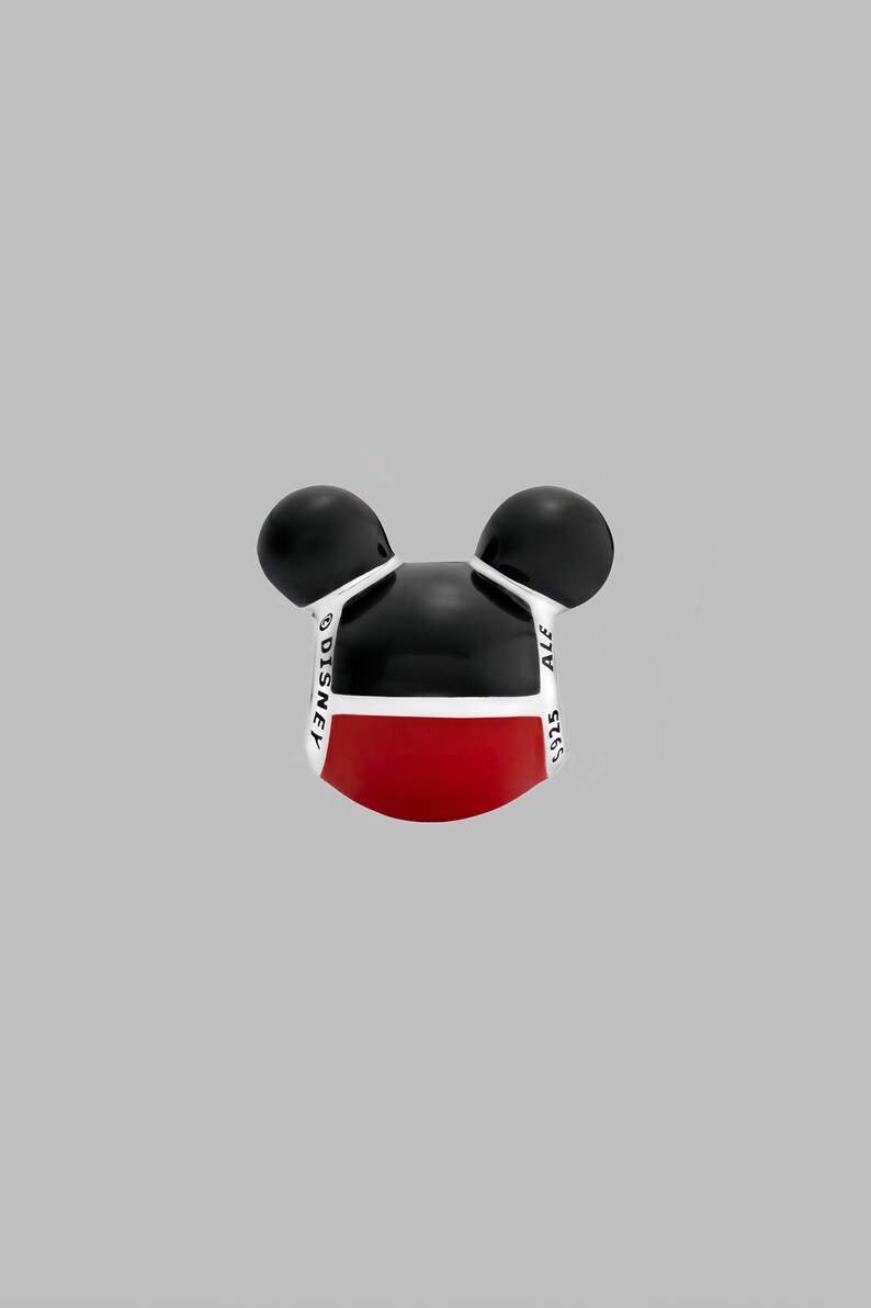 May include: A Mickey Mouse-shaped charm with black ears, a black head, and a red lower face. The charm has white accents and the words "DISNEY" and "ALE 925" printed on it. The charm is set against a gray background.