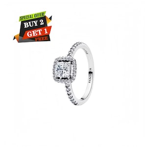 May include: A silver-tone ring featuring a central cushion-cut clear stone surrounded by a halo of smaller clear stones. The band is pavé-set with clear stones and engraved with the text PANDORA and S925 on the inner surface of the band.