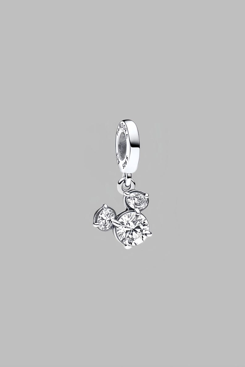 May include: A silver charm featuring three clear cubic zirconia stones, forming a Mickey Mouse head shape. The charm has a loop for attaching to jewellery. The background is a plain grey.