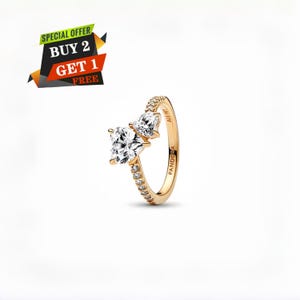 Può includere: Un anello color oro con due pietre preziose trasparenti e scintillanti. Una gemma è a taglio quadrato, l'altra a forma di pera. La fascia è ornata da piccole pietre trasparenti. L'anello reca la scritta "PANDORA". L'immagine include anche il testo: "SPECIAL OFFER BUY 2 GET 1 FREE".