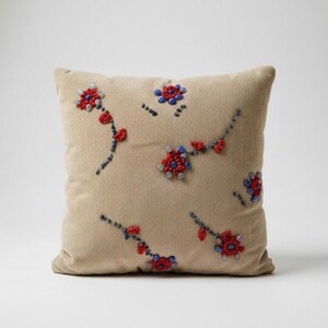 Puede incluir: Cojín cuadrado beige con diseño floral bordado. Las flores son rojas y azules con pequeñas cuentas blancas. El cojín tiene una textura de pana y es un elemento decorativo para el hogar.