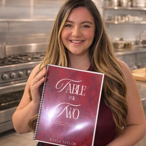 Peut inclure: Un livre de cuisine intitulé "Table for Two" avec une couverture bordeaux et une reliure à spirale blanche. La couverture présente une écriture blanche élégante et le nom de l'auteur, Kayla Taylor. Le livre est présenté devant une cuisine.