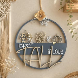 Può includere: Una decorazione da parete circolare blu scuro con la scritta "let's FALL in love" in bianco e tonalità legno chiaro. L'oggetto è decorato con un fiore dorato, una foglia d'acero bianca, un fungo, uno scoiattolo, una ghianda e una zucca.