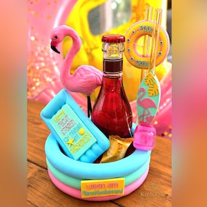 Könnte beinhalten: Eine Miniatur-Pool-Dekoration mit einer rosa Flamingo-Figur, einer Glasflasche mit roter Flüssigkeit, einem gelben Pool-Schwimmring mit der Aufschrift POOL CREW und einem blauen rechteckigen Schwimmkörper mit der Aufschrift BITTE NICHT STÖREN ICH KRIECHE IM PARADIES.