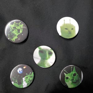 May include: Five circular pinback buttons featuring green alien-themed cat illustrations. One button shows a cat with antennae on a dark background with stars, another a green cat head on white, a green alien, a cat on the moon, and a green cat.