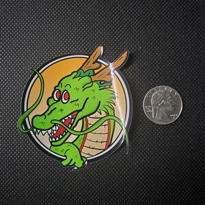 May include: A sticker featuring a green dragon with red eyes and a white-toothed grin, set against a yellow and tan circular background. The dragon has brown antlers and is surrounded by a black border. A US quarter is next to the sticker.