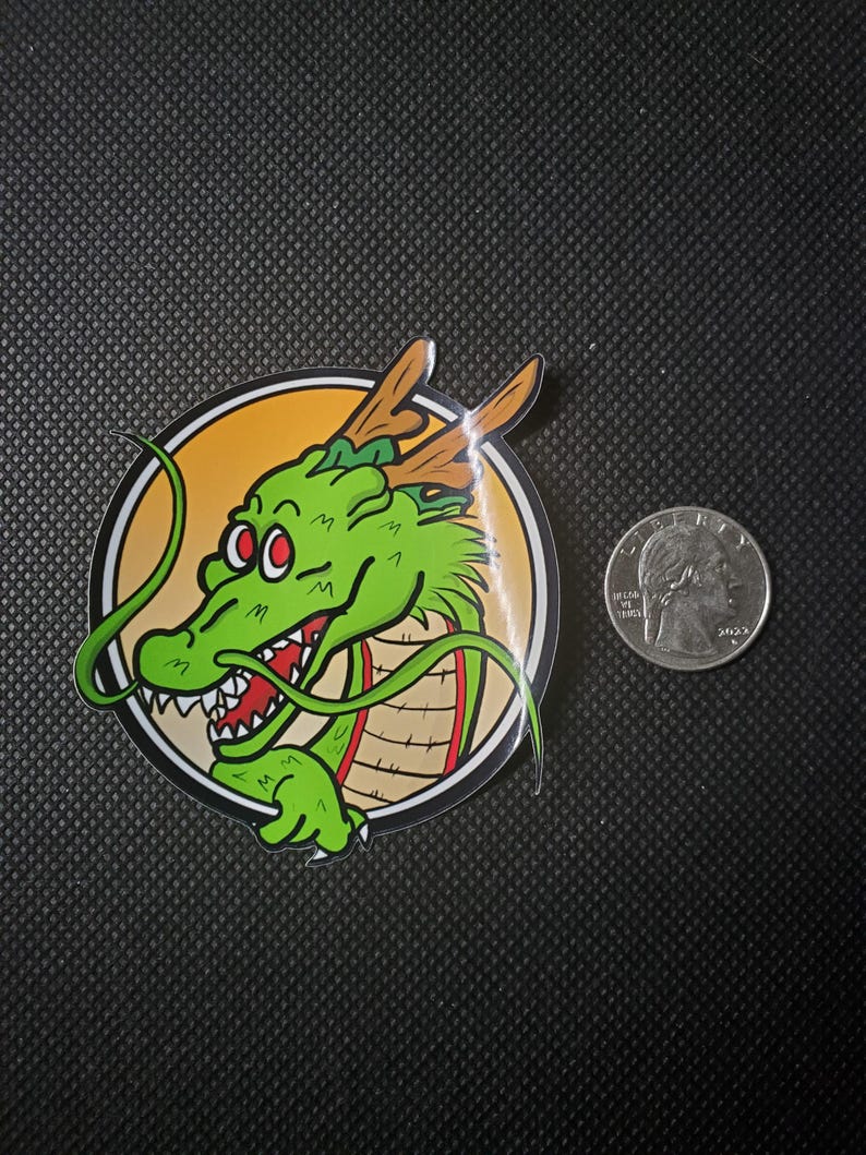 May include: A sticker featuring a green dragon with red eyes and a white-toothed grin, set against a yellow and tan circular background. The dragon has brown antlers and is surrounded by a black border. A US quarter is next to the sticker.