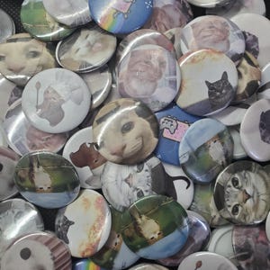 May include: A collection of round pinback buttons featuring various cat images. The buttons display cat faces, Nyan Cat, and other cat-themed designs. The buttons are in shades of white, gray, orange, and black.