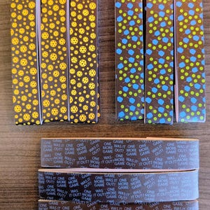May include: Several rectangular boxes with various patterns are displayed. Some boxes feature a pattern of yellow pickleball balls on a dark background. Other boxes have blue and green shapes. The bottom boxes have text that reads "ONE GAME WAS IT OUT? MORE GAME OUT?"