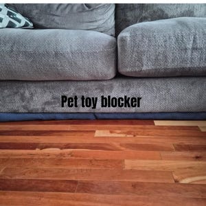 May include: A blue and white patterned pet toy blocker is positioned beneath a grey sofa. The text "Pet toy blocker" is displayed in black. The floor is made of wood.