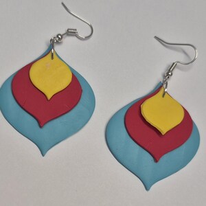 May include: A pair of layered drop earrings featuring three stacked petal-shaped components in bright yellow, red, and light blue. Each earring is suspended from a silver-toned metal hook wire, creating a tiered, teardrop-like silhouette.