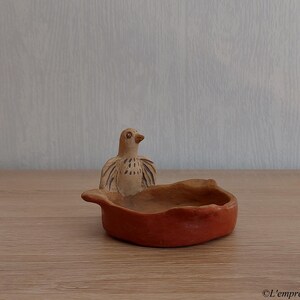 May include: A small, round, terracotta-coloured clay bowl featuring a sculpted bird figure attached to one side. The bird has light brown plumage with dark brown painted details on its wings and face, while the bowl has an uneven, organic rim.