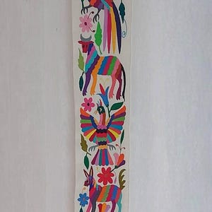 Op de afbeelding: Geborduurde textiel met kleurrijke dieren- en bloemmotieven. Het kunstwerk toont verschillende wezens, waaronder vogels, konijnen en een stier, allemaal in levendige kleuren. Het is een verticaal rechthoek, waarschijnlijk een wandkleed.