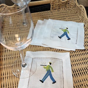 May include: White linen cocktail napkins with embroidered waiter design. The waiter is wearing a green jacket, blue pants, and a black hat. The napkins are on a woven tray with two wine glasses.