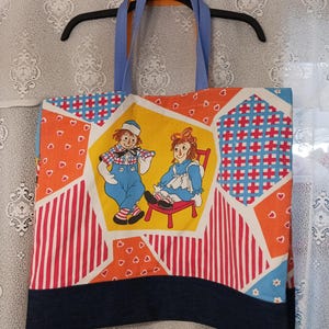 May include: A tote bag featuring a patchwork design with orange, blue, and white patterns, including stripes, hearts, and a central yellow hexagon panel depicting two rag dolls. The bag has blue fabric handles and a dark blue denim-like bottom border.