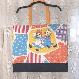 May include: A rectangular tote bag with orange fabric handles featuring a patchwork design of Raggedy Ann and Andy characters, red stripes, blue floral patterns, and orange heart prints, finished with a solid dark blue band at the bottom.