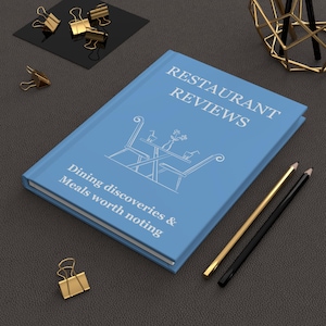 May include: A light blue book titled "Restaurant Reviews" with the subtitle "Dining discoveries & Meals worth noting." The cover features a white line drawing of a restaurant table setting. Gold and black pencils and binder clips are also present.