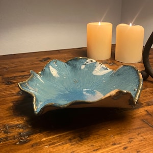 May include: A handmade ceramic bowl with a unique, organic shape and a glossy blue glaze. The rim has a natural, unglazed edge with a touch of gold. Two lit candles are in the background, creating a warm ambiance.