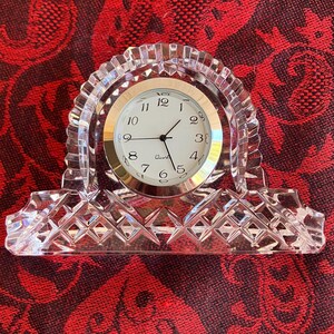May include: A clear crystal quartz clock with a gold-coloured clock face and black hands. The clock has a decorative, arched crystal frame with a diamond-patterned base. The clock face displays the numbers 1 to 12.