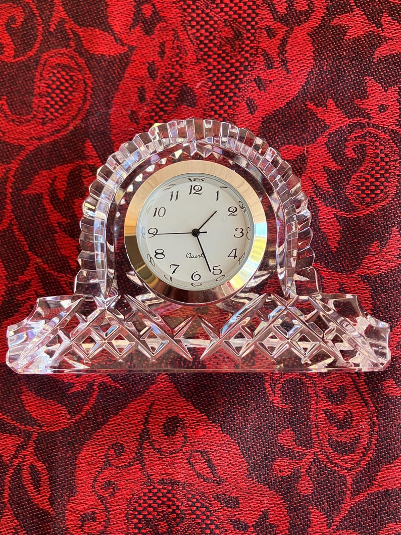 May include: A clear crystal quartz clock with a gold-coloured clock face and black hands. The clock has a decorative, arched crystal frame with a diamond-patterned base. The clock face displays the numbers 1 to 12.