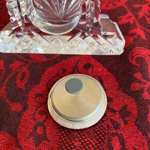 May include: A clear, cut-glass clock with a circular face and a silver-coloured base. A small, round, silver-coloured object with a grey centre is in the foreground. The clock has a decorative, geometric design.