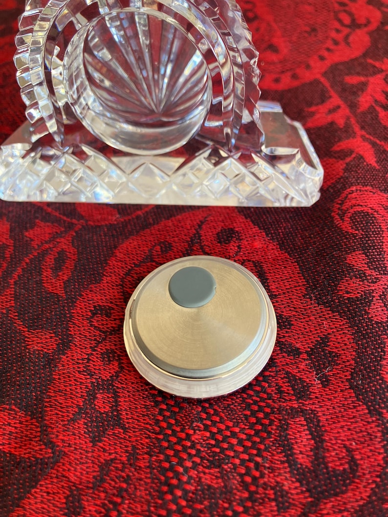 May include: A clear, cut-glass clock with a circular face and a silver-coloured base. A small, round, silver-coloured object with a grey centre is in the foreground. The clock has a decorative, geometric design.