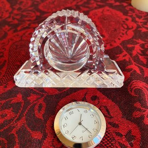 May include: A clear crystal clock with a silver clock face. The clock has a rectangular base and a domed top with intricate cut glass patterns. The clock face has black numerals and hands, with the word "Quartz" printed below the hands.