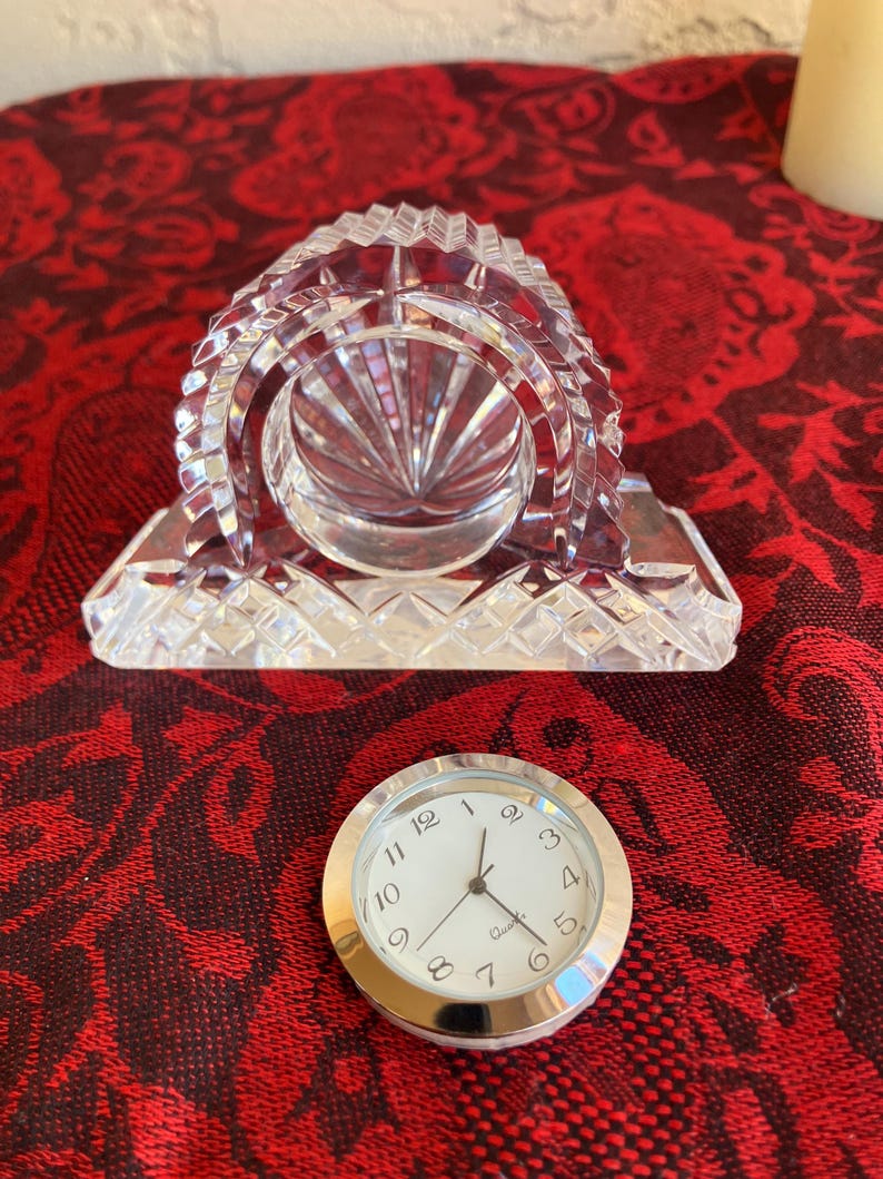 May include: A clear crystal clock with a silver clock face. The clock has a rectangular base and a domed top with intricate cut glass patterns. The clock face has black numerals and hands, with the word "Quartz" printed below the hands.