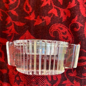 May include: A clear, rectangular crystal candle holder with vertical lines and two small handles. The candle holder is designed to hold a single candle. The object is on a red patterned surface.