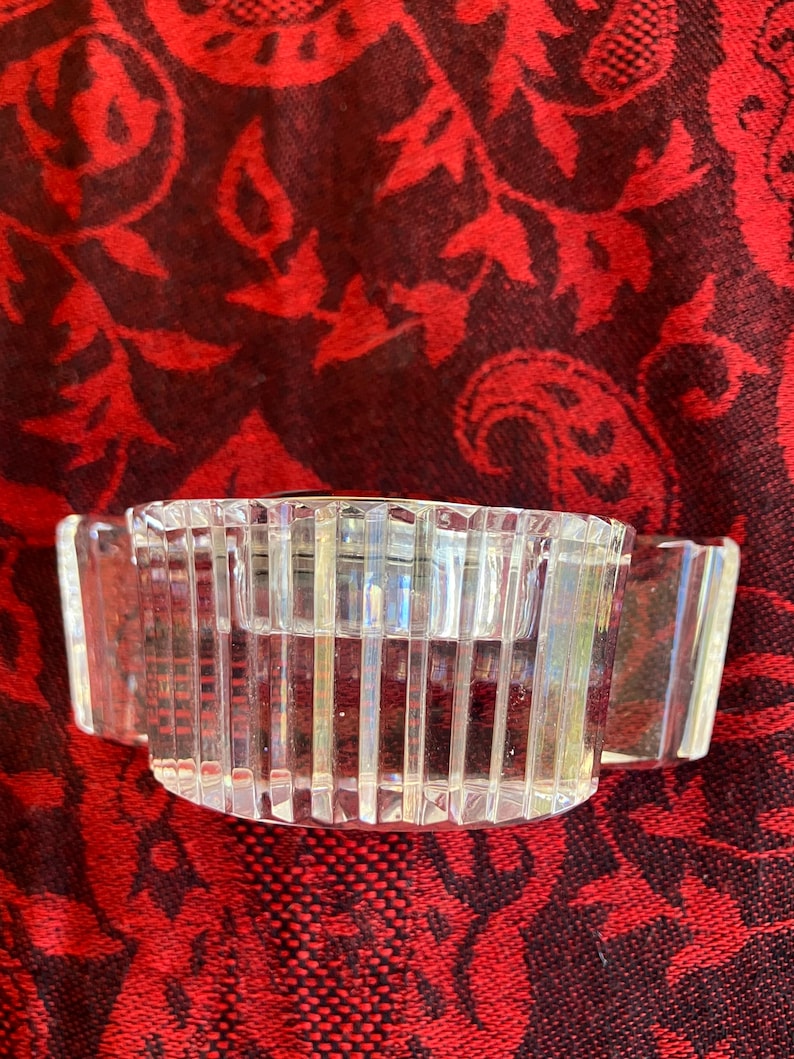 May include: A clear, rectangular crystal candle holder with vertical lines and two small handles. The candle holder is designed to hold a single candle. The object is on a red patterned surface.