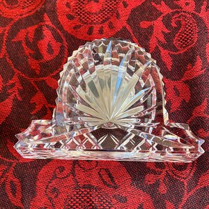 May include: A clear, cut-glass napkin holder with a fan-shaped design. The holder has a rectangular base with decorative edges and a central, arched section. The glass reflects light, creating a sparkling effect. The napkin holder is likely vintage.