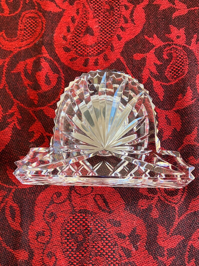 May include: A clear, cut-glass napkin holder with a fan-shaped design. The holder has a rectangular base with decorative edges and a central, arched section. The glass reflects light, creating a sparkling effect. The napkin holder is likely vintage.