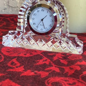 May include: A clear crystal quartz clock with a white face and black numerals. The clock has a silver bezel and is set in a decorative crystal frame. The base of the clock has a diamond pattern.