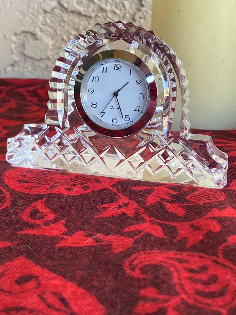 May include: A clear crystal quartz clock with a white face and black numerals. The clock has a silver bezel and is set in a decorative crystal frame. The base of the clock has a diamond pattern.