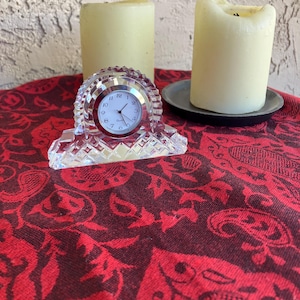 May include: A clear crystal clock with a round face, set on a red patterned fabric. Two cream-coloured candles are in the background, one on a dark grey plate. The clock has a silver-coloured frame and black numbers.
