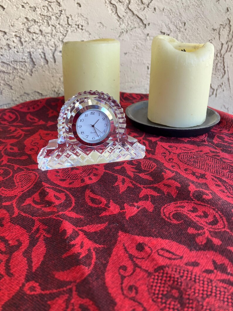 May include: A clear crystal clock with a round face, set on a red patterned fabric. Two cream-coloured candles are in the background, one on a dark grey plate. The clock has a silver-coloured frame and black numbers.