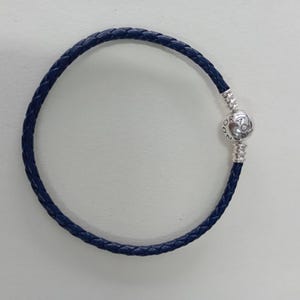 Könnte beinhalten: Ein marineblaues geflochtenes Lederarmband mit einem silbernen Verschluss. Der Verschluss ist eine runde Perle mit dem eingravierten Markennamen. Das Armband ist ein schlichtes, elegantes Schmuckstück.
