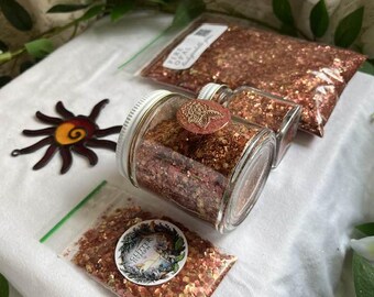 May include: An assortment of glitter in various containers. There are two small glass jars with lids, a clear plastic bag, and a larger plastic bag, all filled with glitter. A decorative metal sun and greenery are also present.