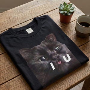 以下が含まれることがあります： 黒いTシャツ。黒猫の顔のクローズアップと白い文字「I U」がプリントされています。猫は大きな目とピンクの舌をしています。Tシャツは木製のテーブルの上に折りたたまれています。
