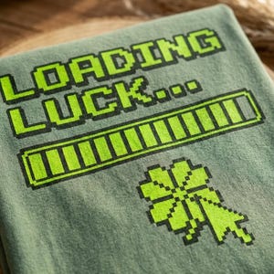 Könnte beinhalten: Ein grünes T-Shirt mit dem Text "LOADING LUCK..." in einem pixeligen Stil. Darunter befindet sich ein Ladebalken und ein pixeliges vierblättriges Kleeblatt. Das Design ist in leuchtendem Grün mit schwarzen Umrissen.