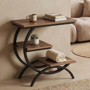 May include: A three-tiered side table with a dark brown wood finish and black metal frame. The top shelf holds a mug and books, the middle shelf has a potted succulent, and the bottom shelf has a rolled-up beige blanket.