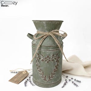 Op de afbeelding: Een verweerde groene metalen vaas met een hart- en bloemmotief, vastgebonden met touw. De vaas heeft twee handvatten en een rustieke uitstraling. Een houten label en lavendel takjes dragen bij aan de landelijke stijl.