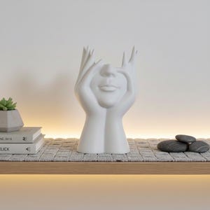 May include: A white ceramic vase shaped like a face with hands framing the eyes. The vase has a smooth finish and is displayed on a wooden shelf with books, a small succulent, and decorative stones. The design is modern and artistic.